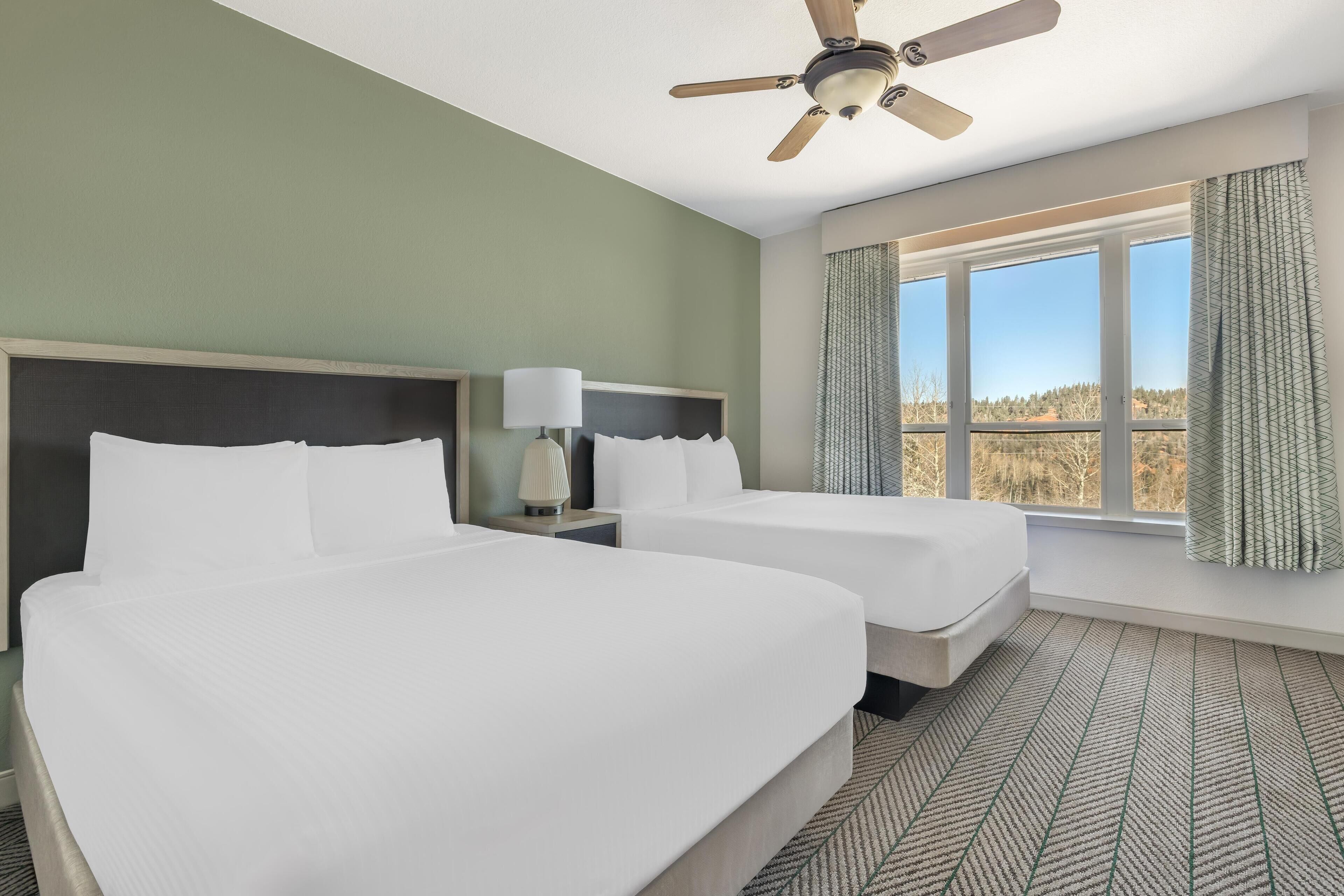 A hotel room with two beds, a ceiling fan, a window with curtains, and a view of the outdoors.
