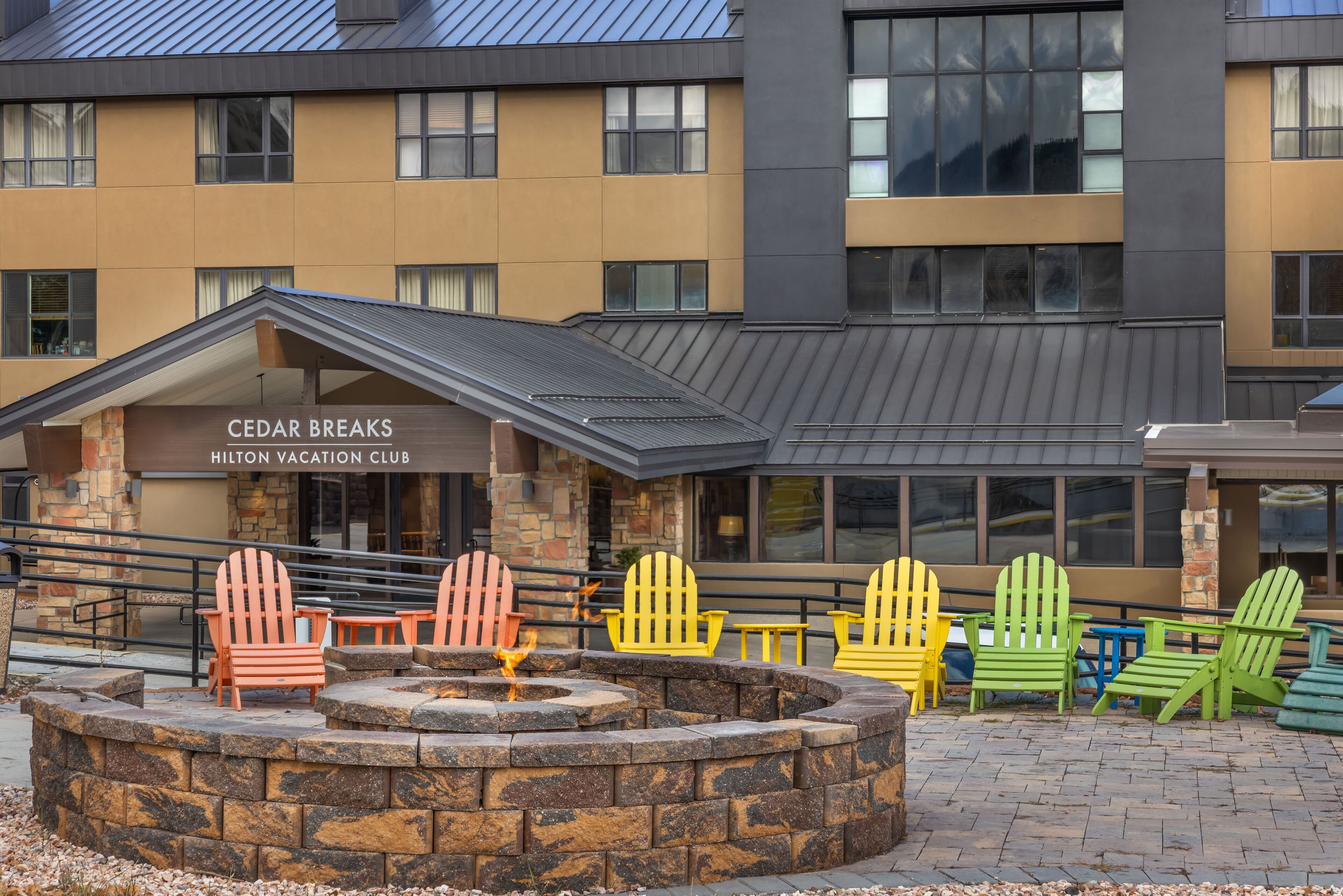 A hotel exterior with a stone fireplace, colorful Adirondack chairs, and a sign for Cedar Breaks Hilton Vacation Club.