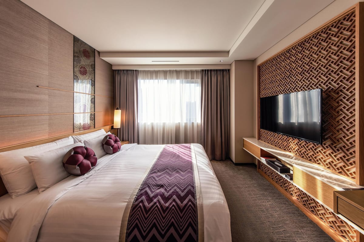 Quarto de hotel moderno com uma cama grande, uma TV de tela plana fixada em um painel de madeira e vista para a cidade pela janela.