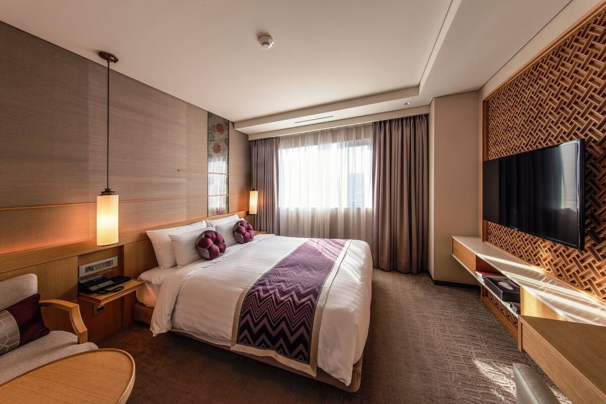 Quarto de hotel com uma cama grande, televisão fixada na parede, secretária com abajur e janela com cortinas.