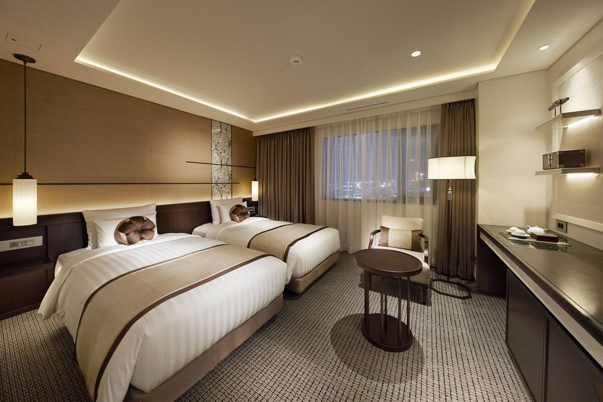 Quarto de hotel com duas camas, uma mesa redonda pequena, uma escrivaninha com cadeira, uma janela grande com cortinas e vista para a cidade.