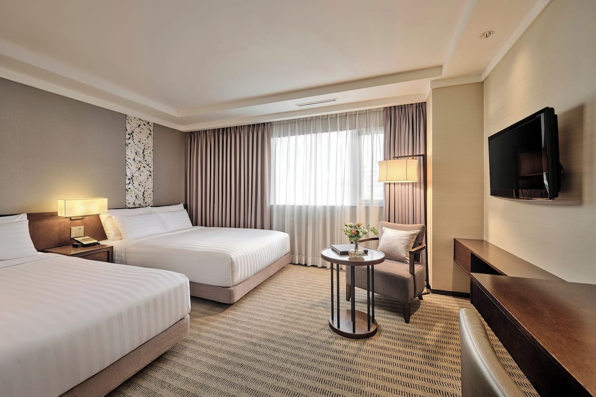 Quarto de hotel com duas camas, uma TV, uma mesinha de cabeceira com abajur e uma janela grande com cortinas.