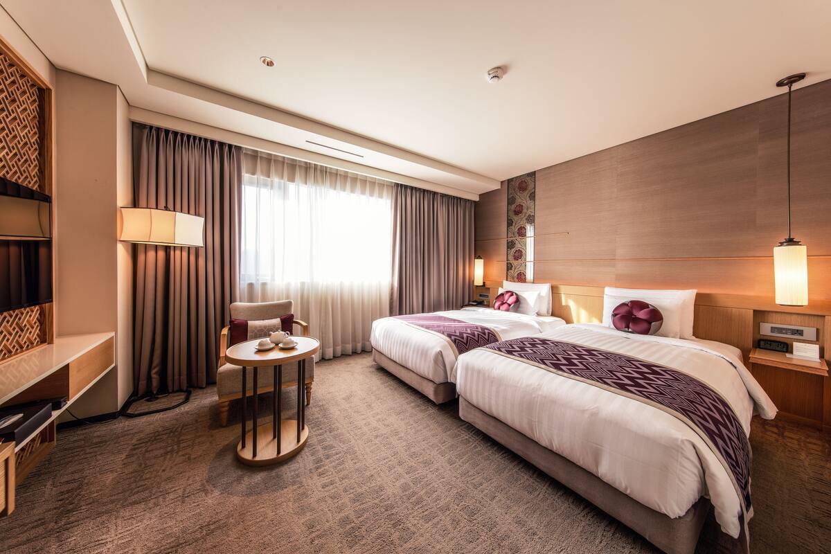 Quarto de hotel com duas camas, uma televisão, uma mesinha e um sofá.
