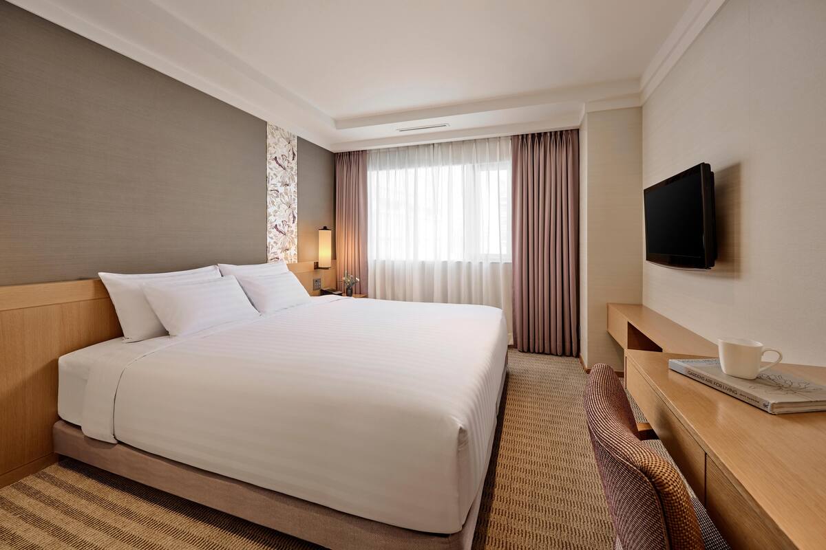 Quarto de hotel com uma cama grande, uma mesa de cabeceira com abajur, uma TV na parede e uma janela com cortinas.