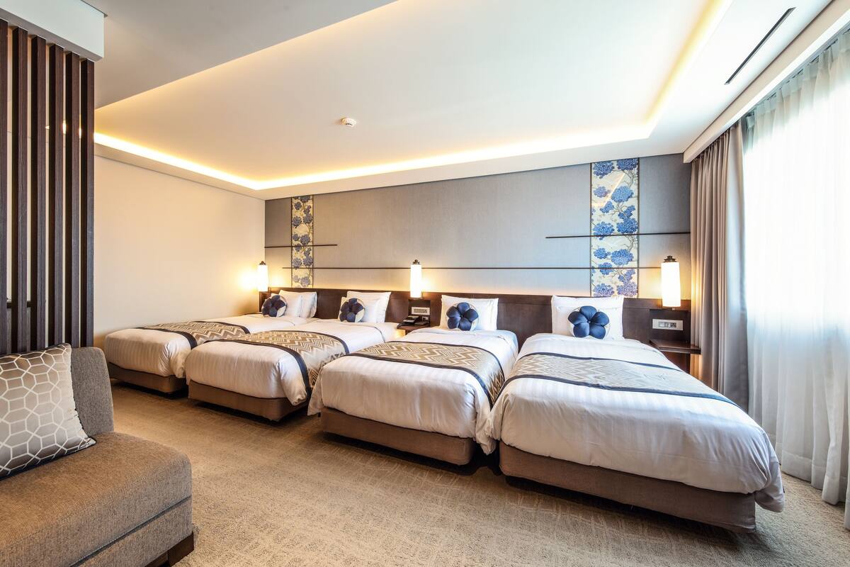 Quarto de hotel com quatro camas dispostas em linha, um sofá e uma parede grande com painéis decorativos.