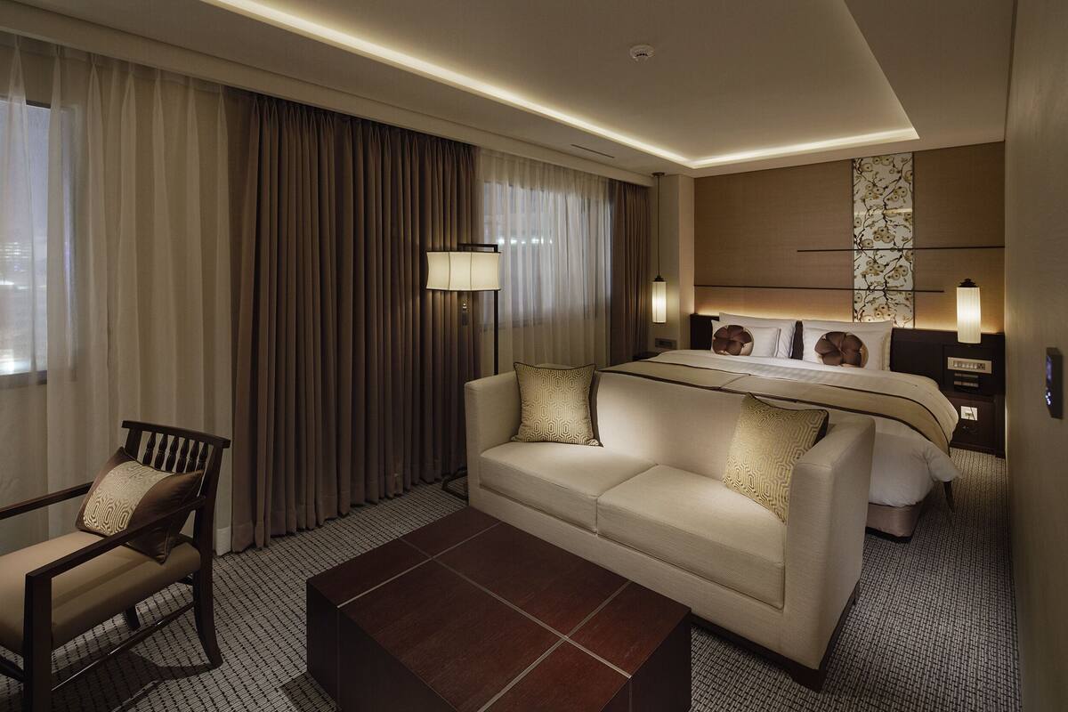 Quarto de hotel com sofá, cadeira, cama, mesa de cabeceira e abajur.