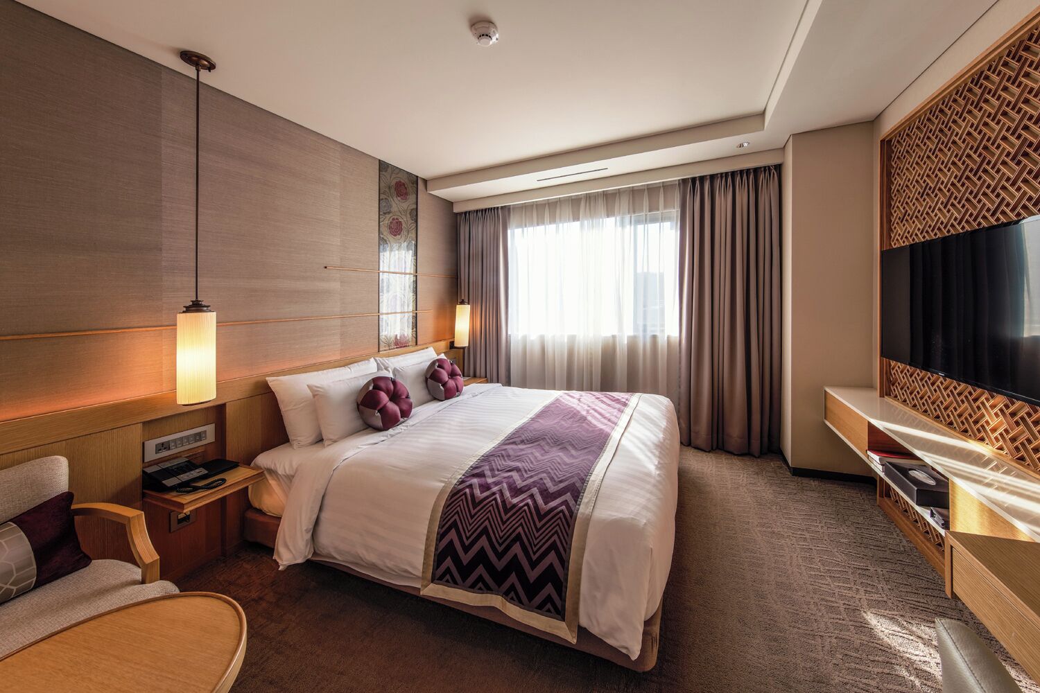 A hotel room with a large bed, bedside tables, a desk, a chair, a TV, and a window with curtains.