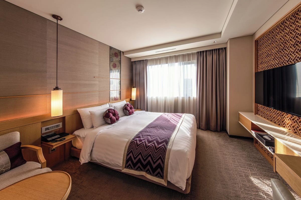 A hotel room with a large bed, bedside tables, a desk, a chair, a TV, and a window with curtains.