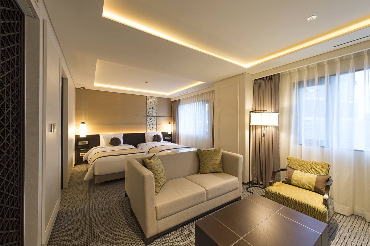 Quarto de hotel com cama, sofá, poltrona, mesa de centro e luminária.
