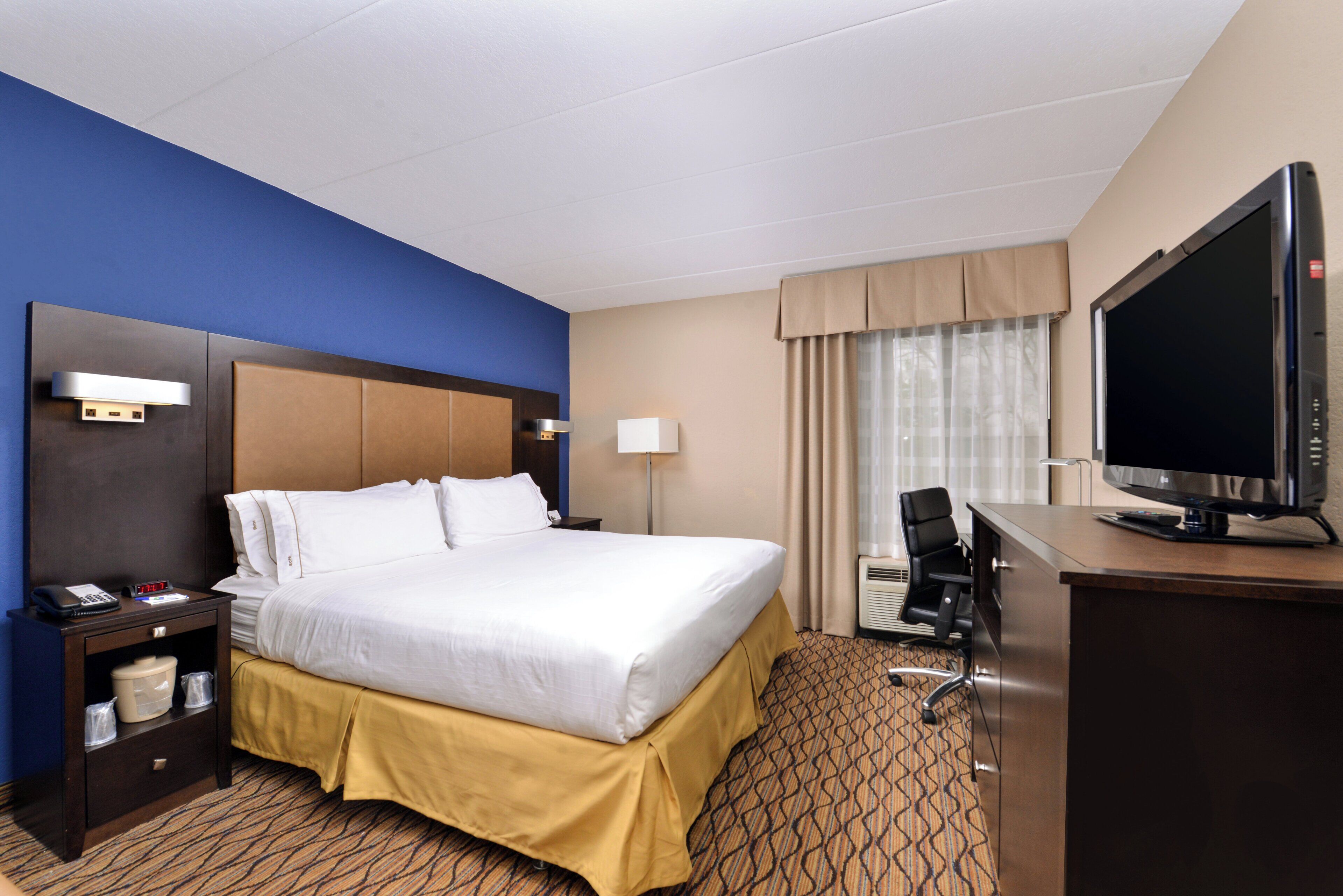 A hotel room with a bed, a desk with a TV, a chair, a nightstand, and a lamp.