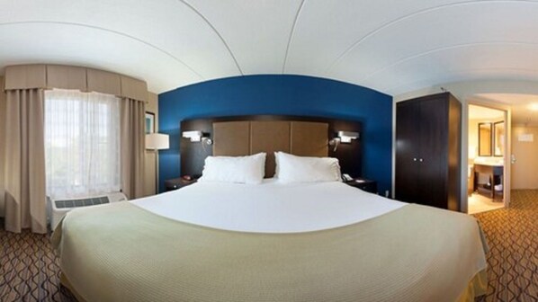 Een groot bed met witte lakens in een hotelkamer met een blauwe accentmuur, een bureau met een lamp en een deur naar een badkamer.