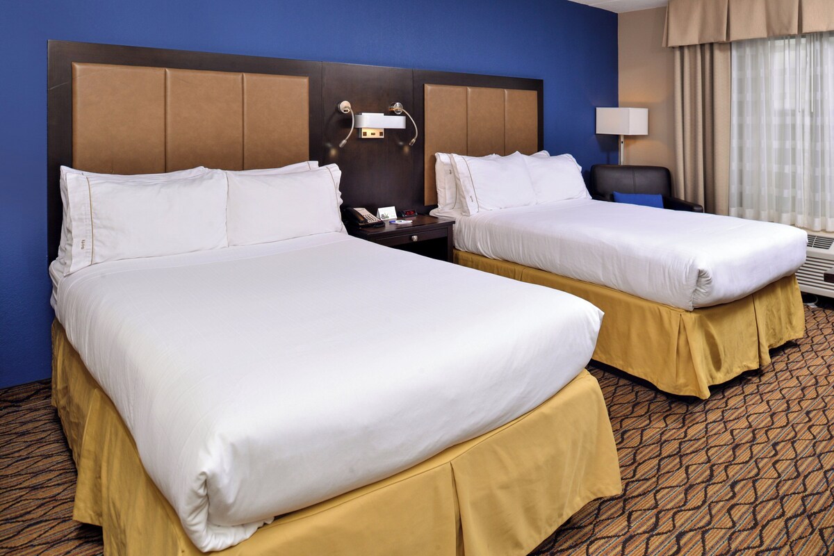 A hotel room with two beds, a nightstand, a lamp, and a window with curtains.