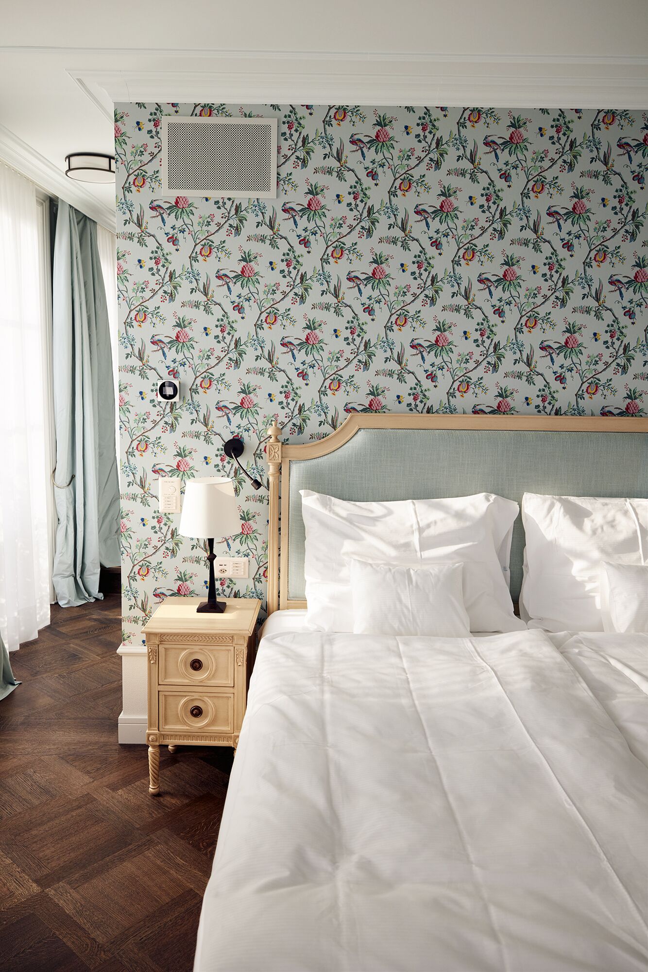 A neatly made bed with white linens, a wooden nightstand with a lamp, and a floral wallpaper pattern.