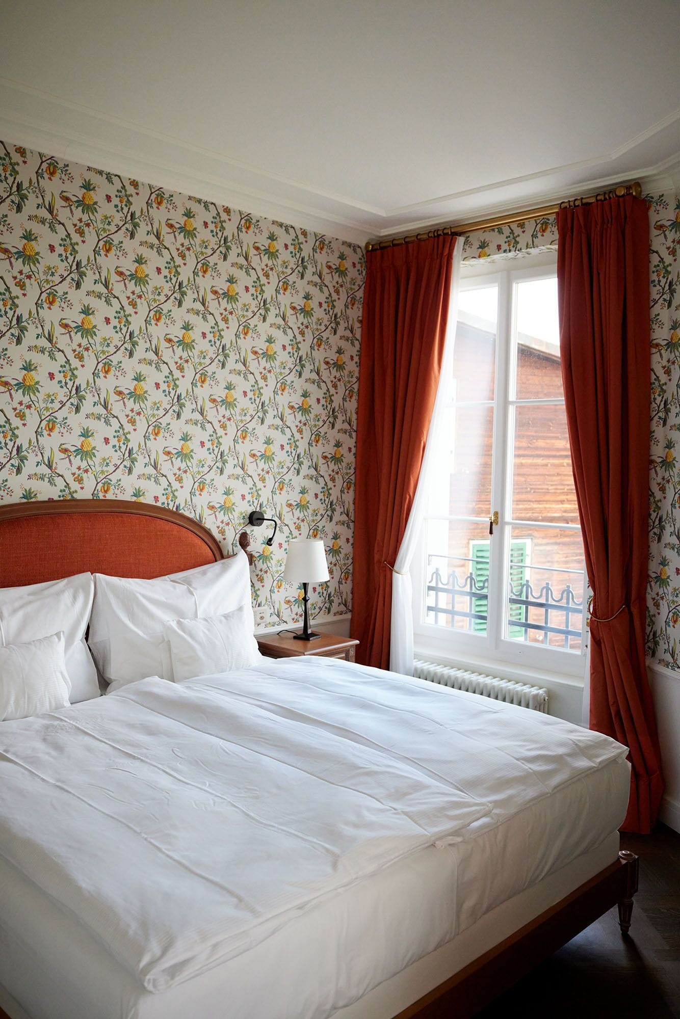 A neatly made bed in a room with floral wallpaper, a nightstand with a lamp, and a window with red curtains.