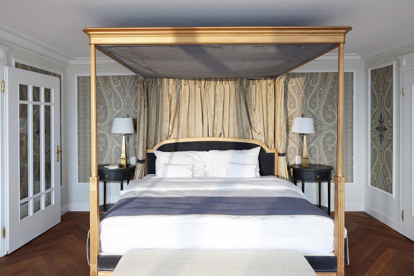 A four-poster bed with a canopy, two bedside tables with lamps, patterned wallpaper, and a wooden floor.