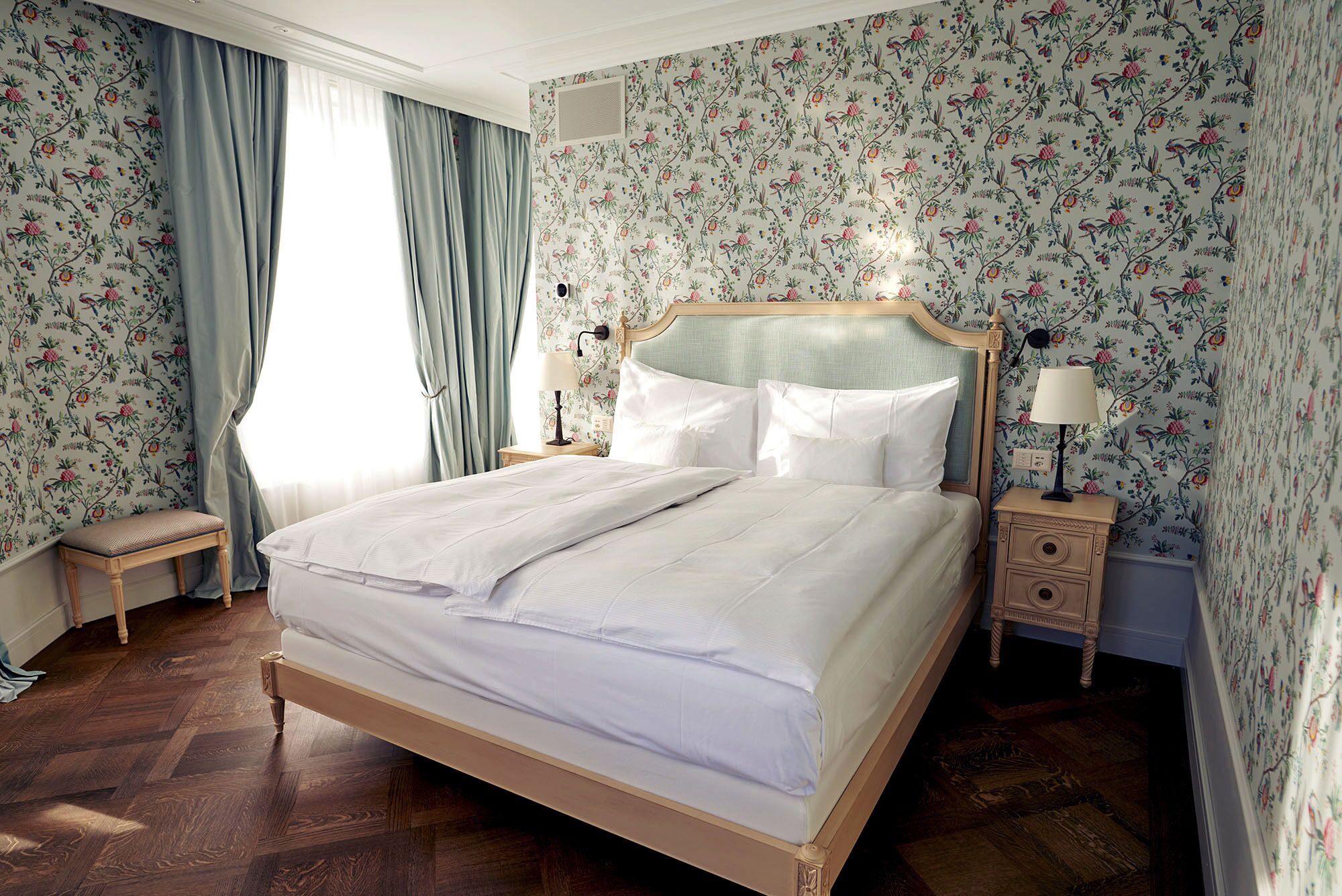 A neatly made bed in a room with floral wallpaper, a wooden floor, and a nightstand with a lamp.