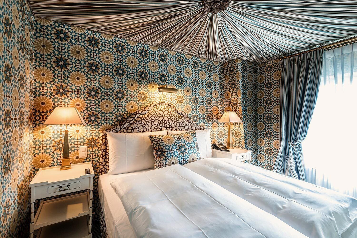 A hotel room with a patterned wall, a bed with white linens, two bedside tables with lamps, and a large window with curtains.
