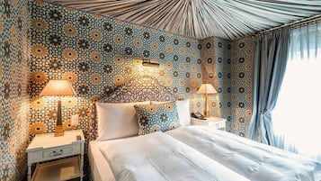 Une chambre d’hôtel avec un mur à motifs, un lit recouvert de draps blancs, deux tables de chevet avec des lampes et une grande fenêtre avec des rideaux.