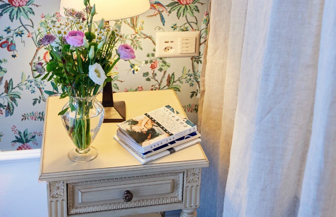 Ein Nachttisch mit Blumen in einer Vase, einer Lampe und einem Stapel Bücher.