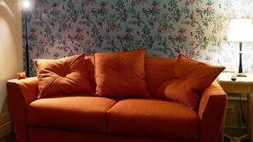 Quarto com sofá de frente para um papel de parede floral, luminária em mesa lateral e tomada visível.