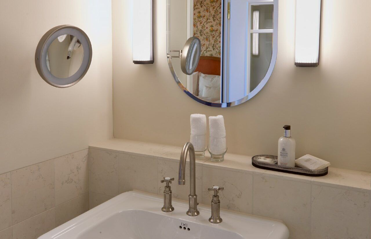 A bathroom with a bathtub, two faucets, a round mirror, and a shelf with toiletries.