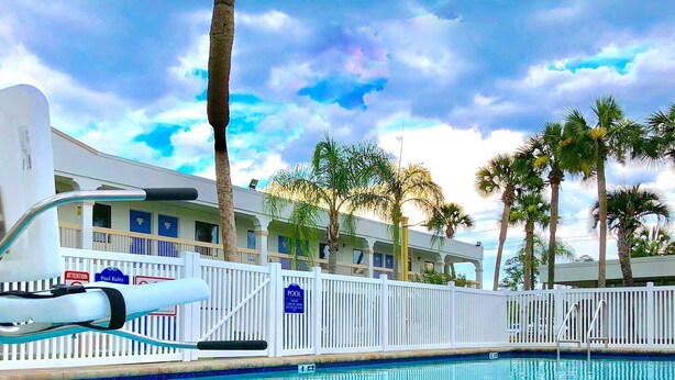 A swimming pool with a white fence, palm trees, and a building with balconies in the background. - Motel 6 Starke, FL (Starke)