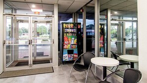 A small indoor seating area with a vending machine stocked with snacks and drinks, and a glass door entrance. - Motel 6 Starke, FL (Starke)