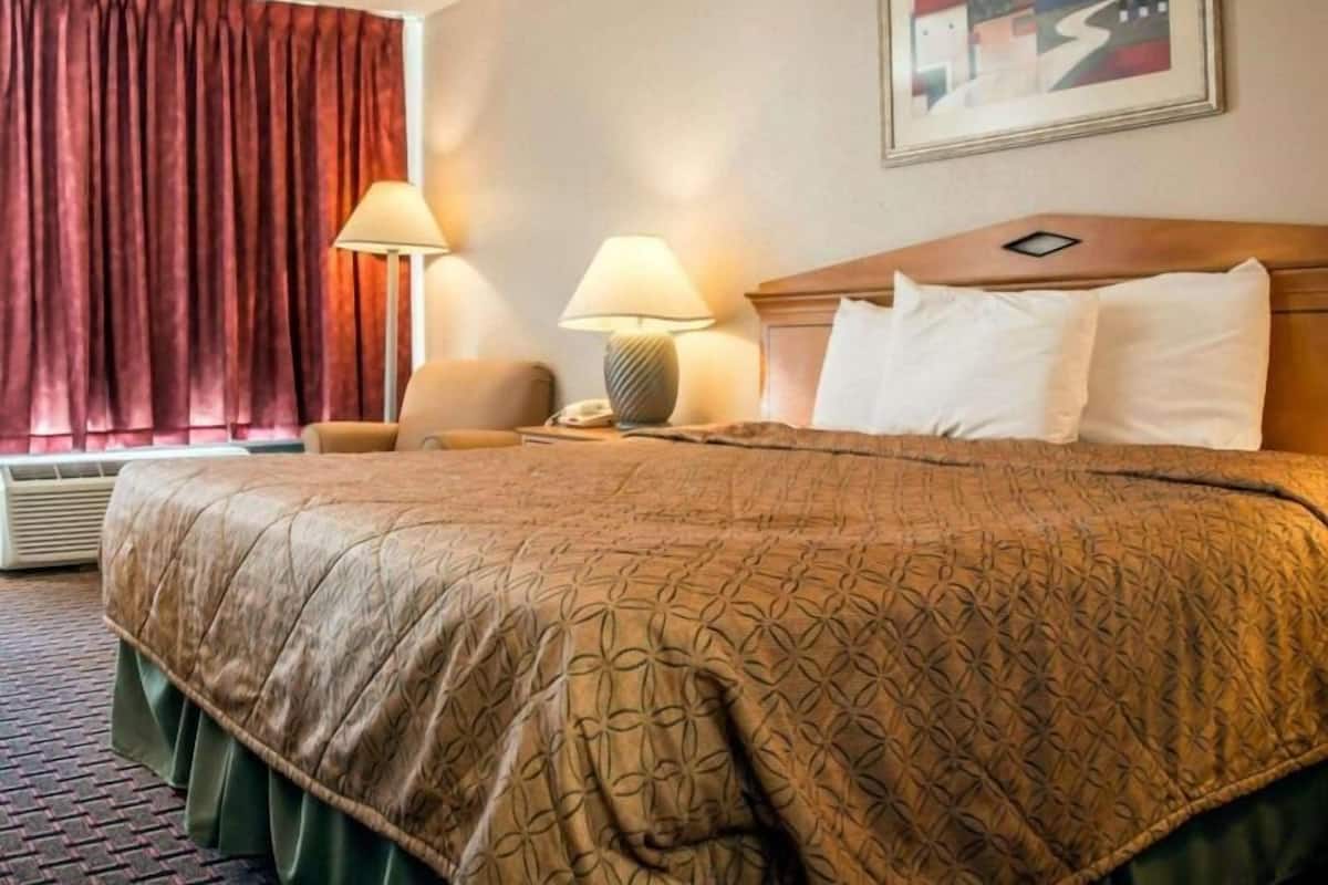 A hotel room with a large bed, two bedside lamps, a chair, a painting on the wall, and a window with curtains.