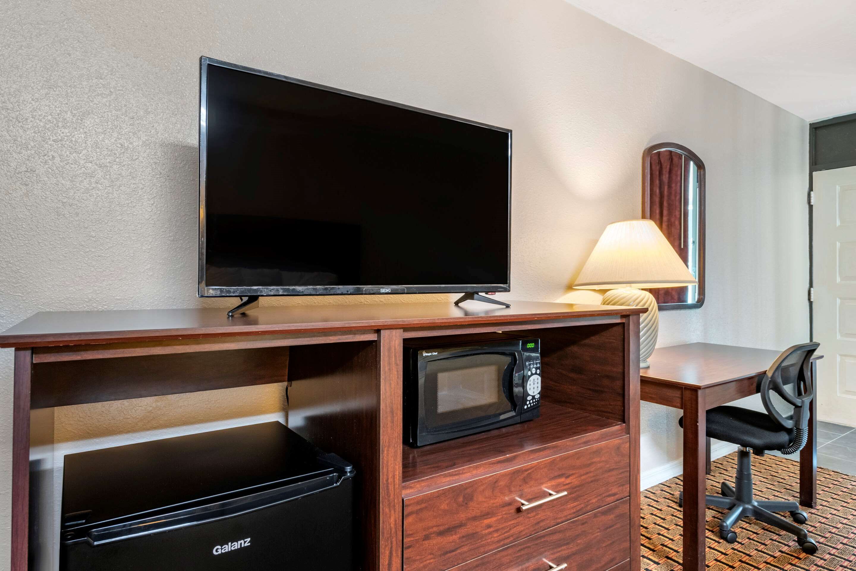 A hotel room with a flat-screen TV, a microwave, a mini fridge, a desk, a chair, and a lamp.