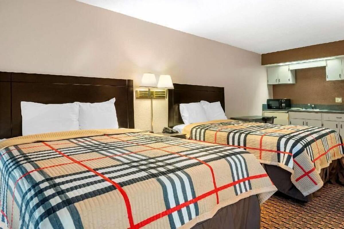 A hotel room with two beds, a nightstand, a lamp, a microwave, and a kitchenette.