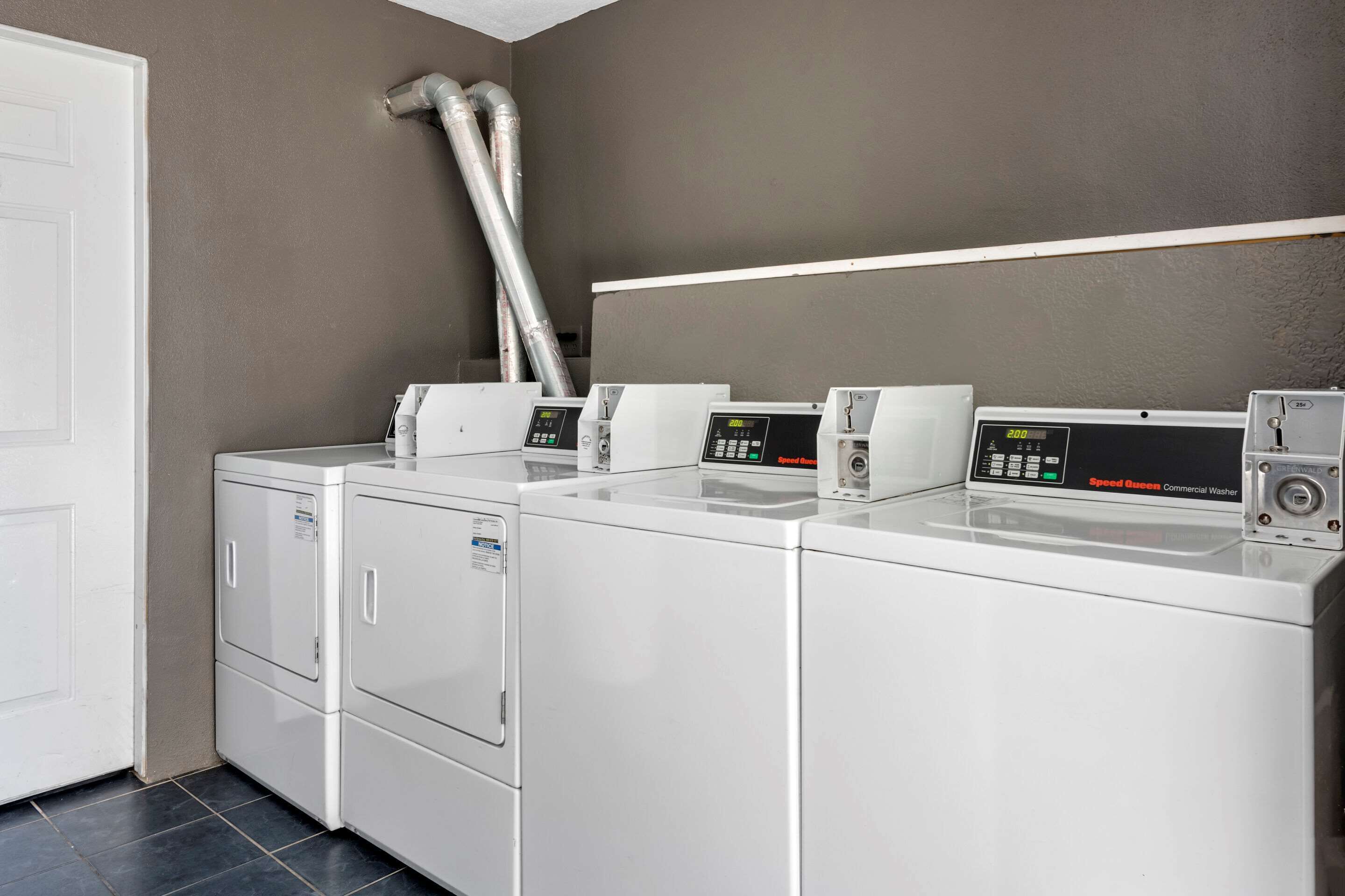A laundry area with four commercial washers and dryers, a white door, and a vent on the wall.