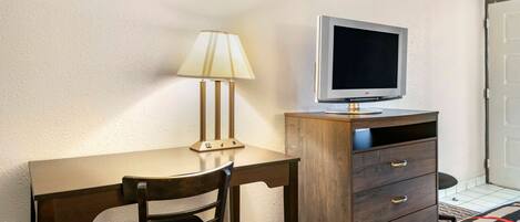 Una habitación con un escritorio de madera, una silla, una lámpara, un televisor sobre un mueble de madera y una cama con colcha a cuadros.