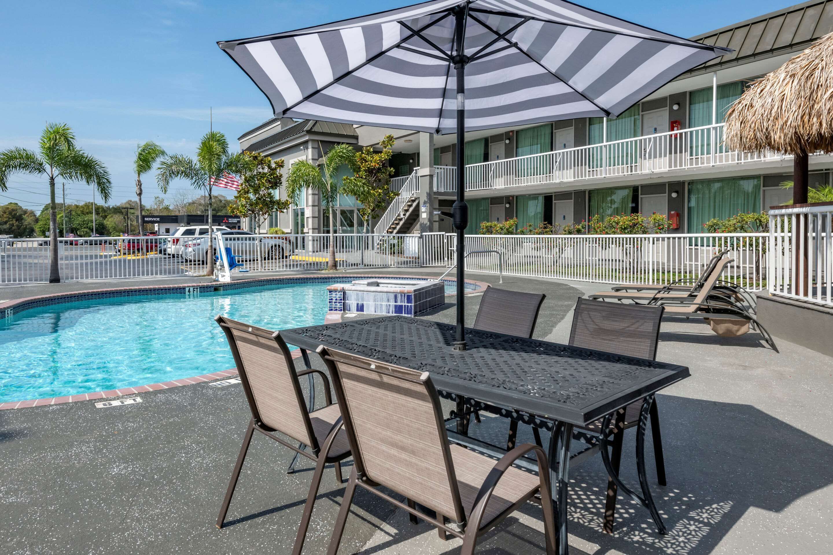 A poolside area with a table and chairs, a striped umbrella, and a building with balconies and a flag.
