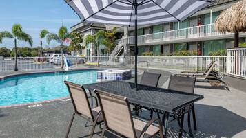 A poolside area with a table and chairs, a striped umbrella, and a building with balconies and a flag.
