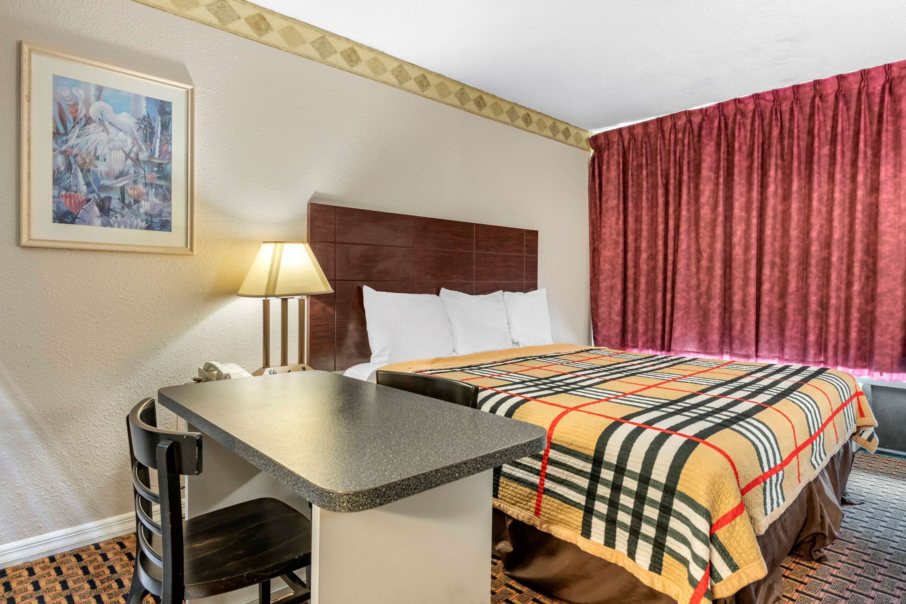 Habitación de hotel con cama, escritorio, silla, lámpara y un cuadro en la pared.