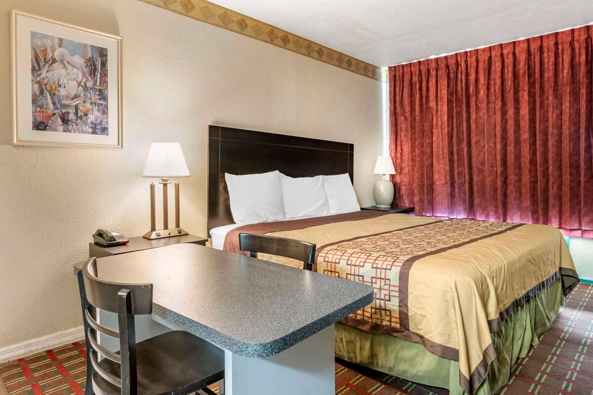 A hotel room with a bed, a desk, two chairs, a telephone, and a painting on the wall.