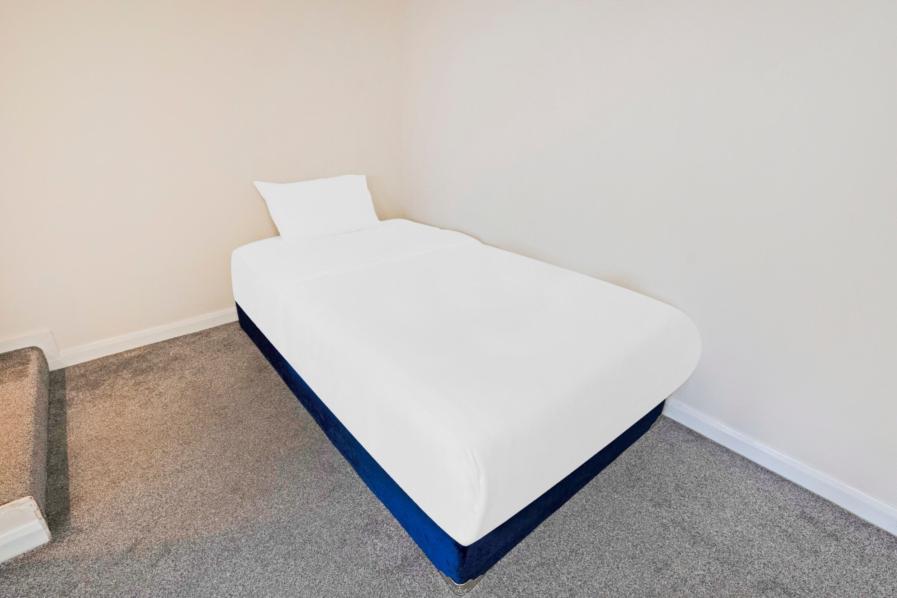 A single bed with a white mattress and blue bed skirt in a room with beige walls and carpeted flooring.