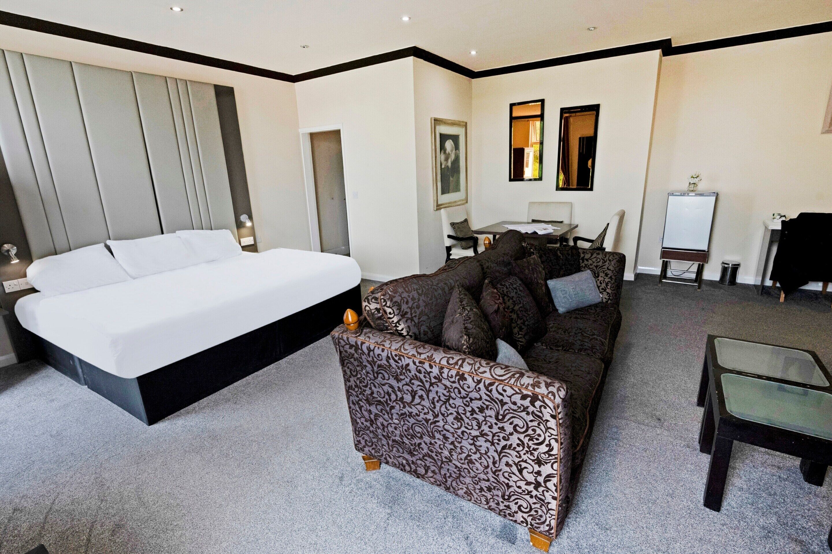 A modern hotel room with a large bed, a patterned sofa, a glass coffee table, and a desk with a chair.