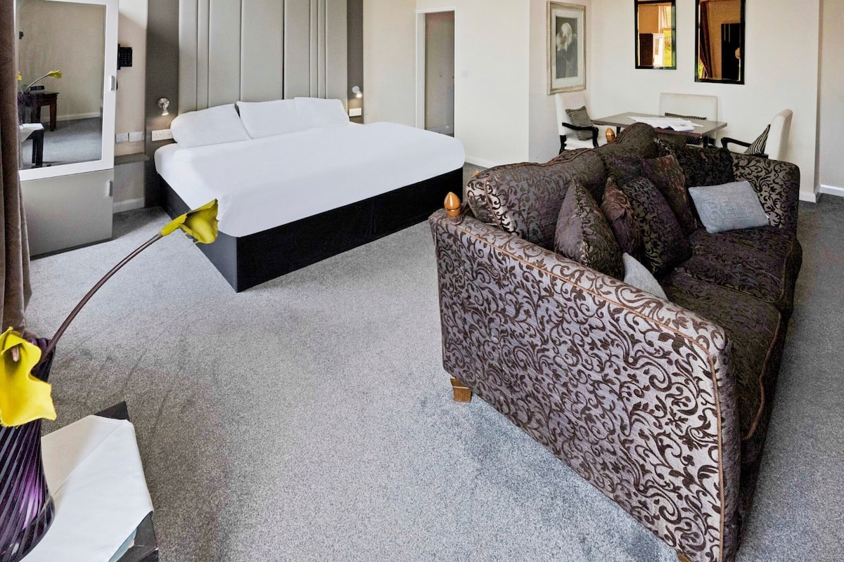 Ett modernt hotellrum med en stor säng, en mönstrad soffa, ett skrivbord och en spegel.