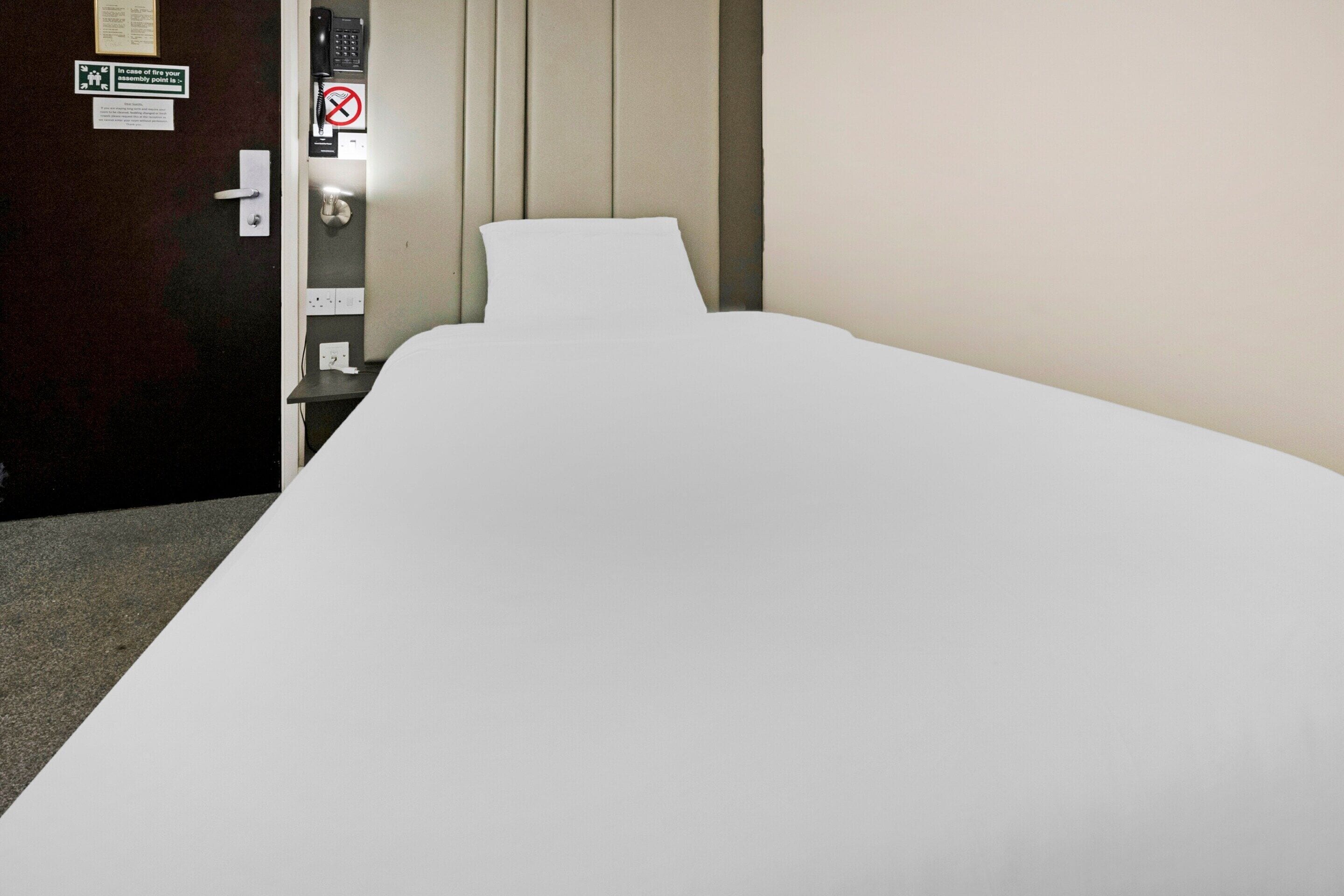 A single bed in a hotel room with a wooden door, a no-smoking sign, and a light switch on the wall.