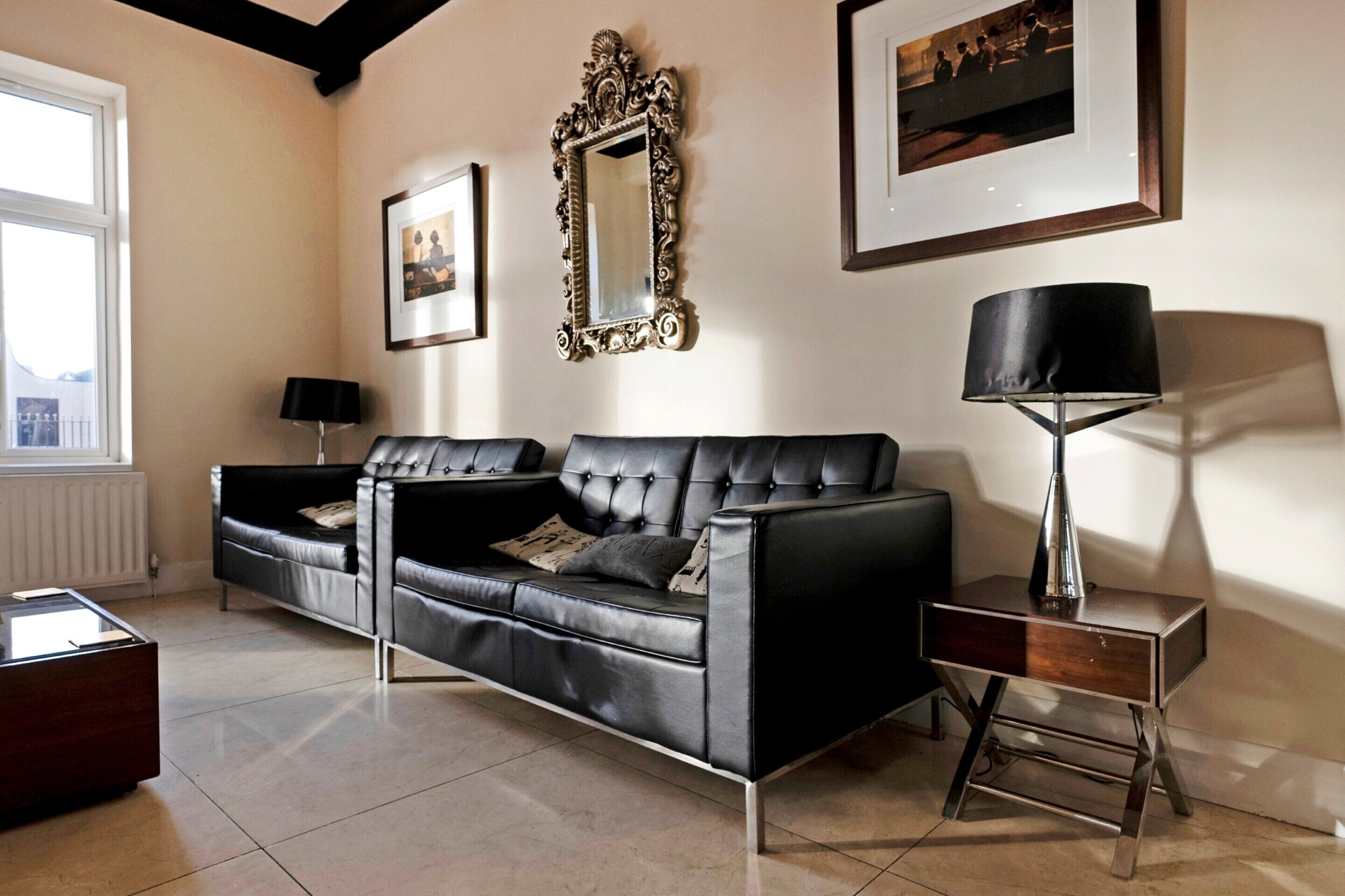 A modern living room with a black leather sofa, a side table with a lamp, and framed pictures on the wall.