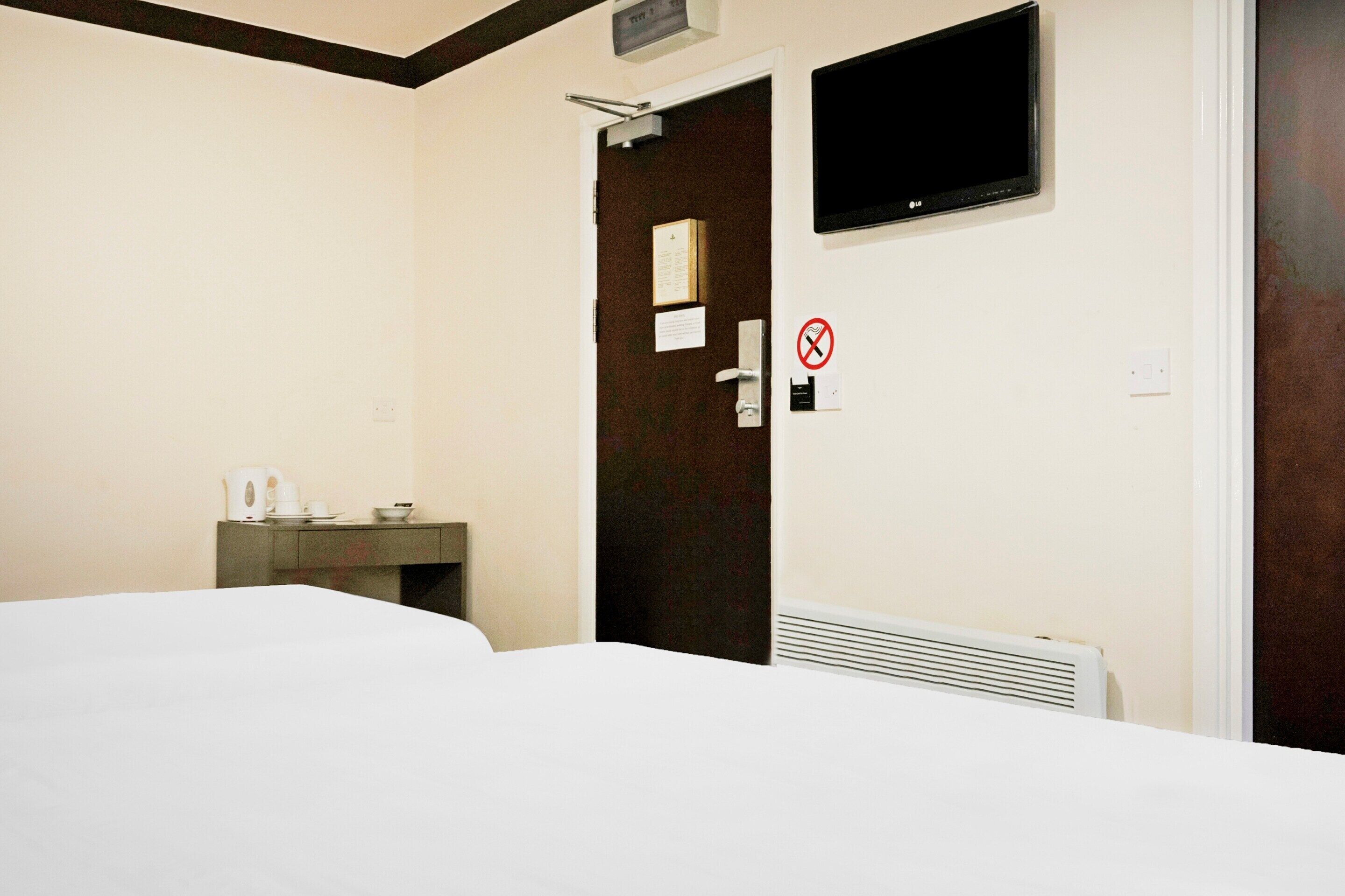 A hotel room with a bed, a small desk with a cup and a bowl, a television mounted on the wall, and a door with a no-smoking sign.