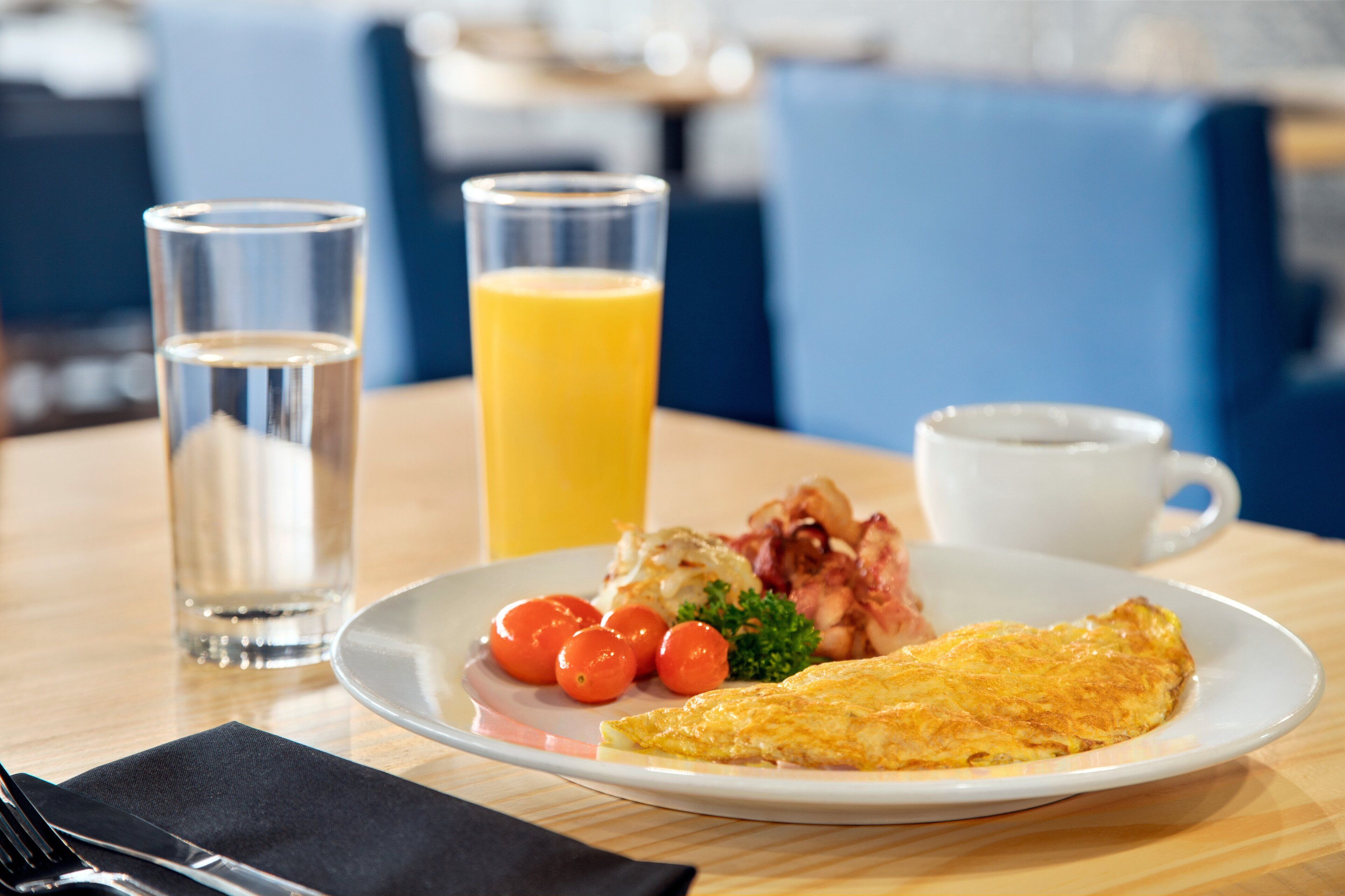 En tallerken med æg, tomater og bacon, et glas vand og et glas appelsinjuice på et træbord.