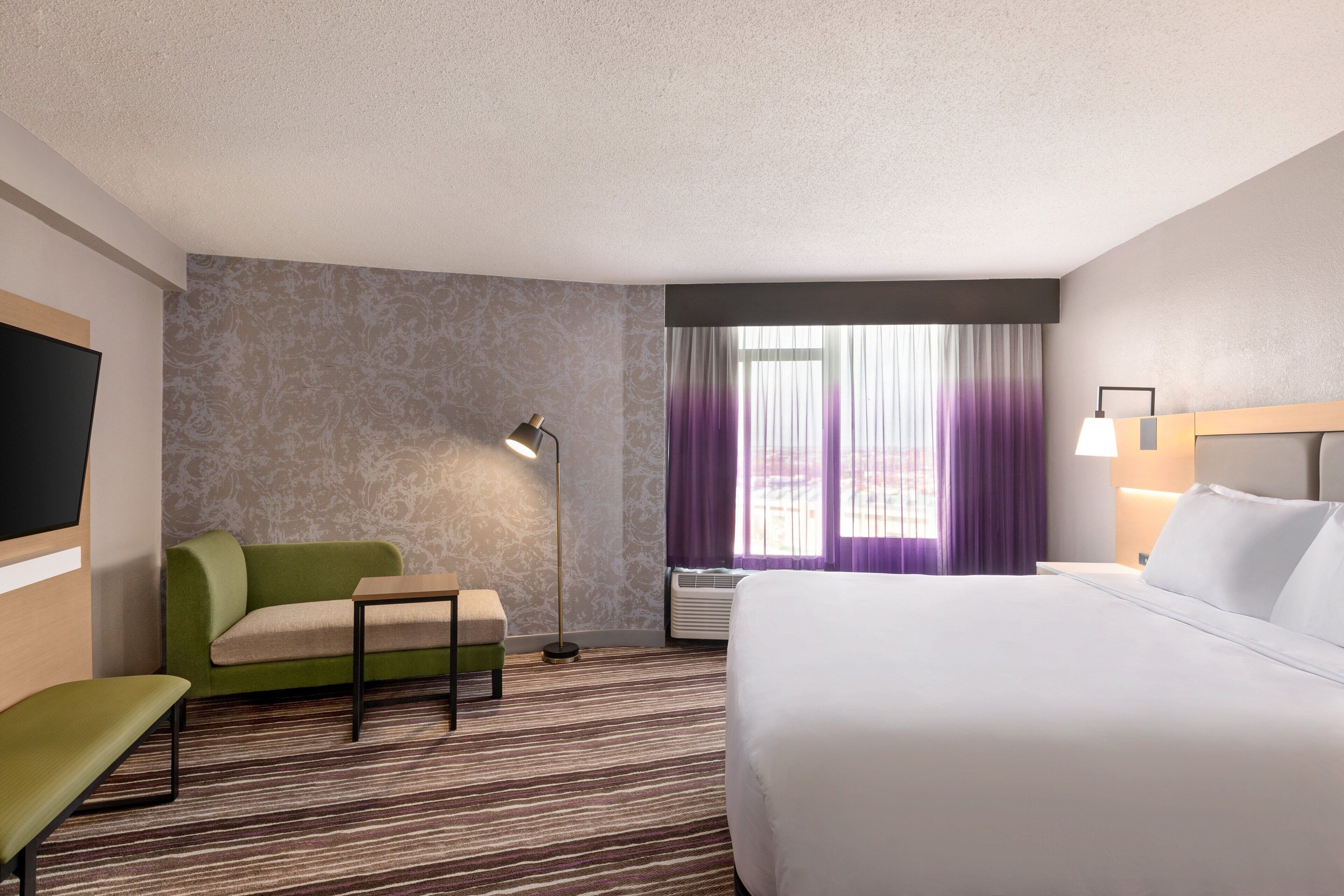 A hotel room with a large bed, a green sofa, a small table, a floor lamp, and a TV.