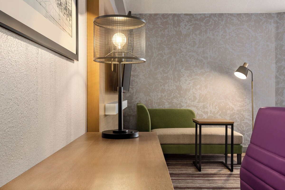 A room with a wooden table, a green sofa, a purple chair, a floor lamp, and a wall-mounted lamp.