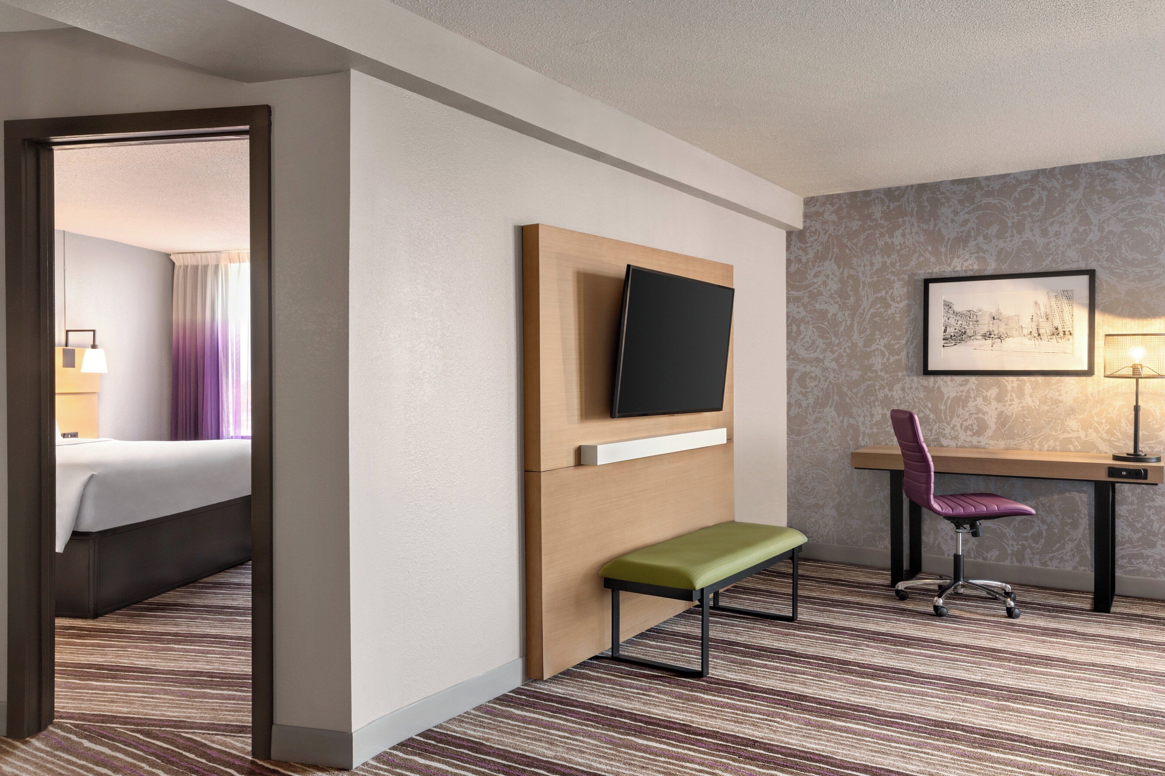 A hotel room with a bed, a desk with a chair, a television, a framed picture on the wall, and a carpeted floor.