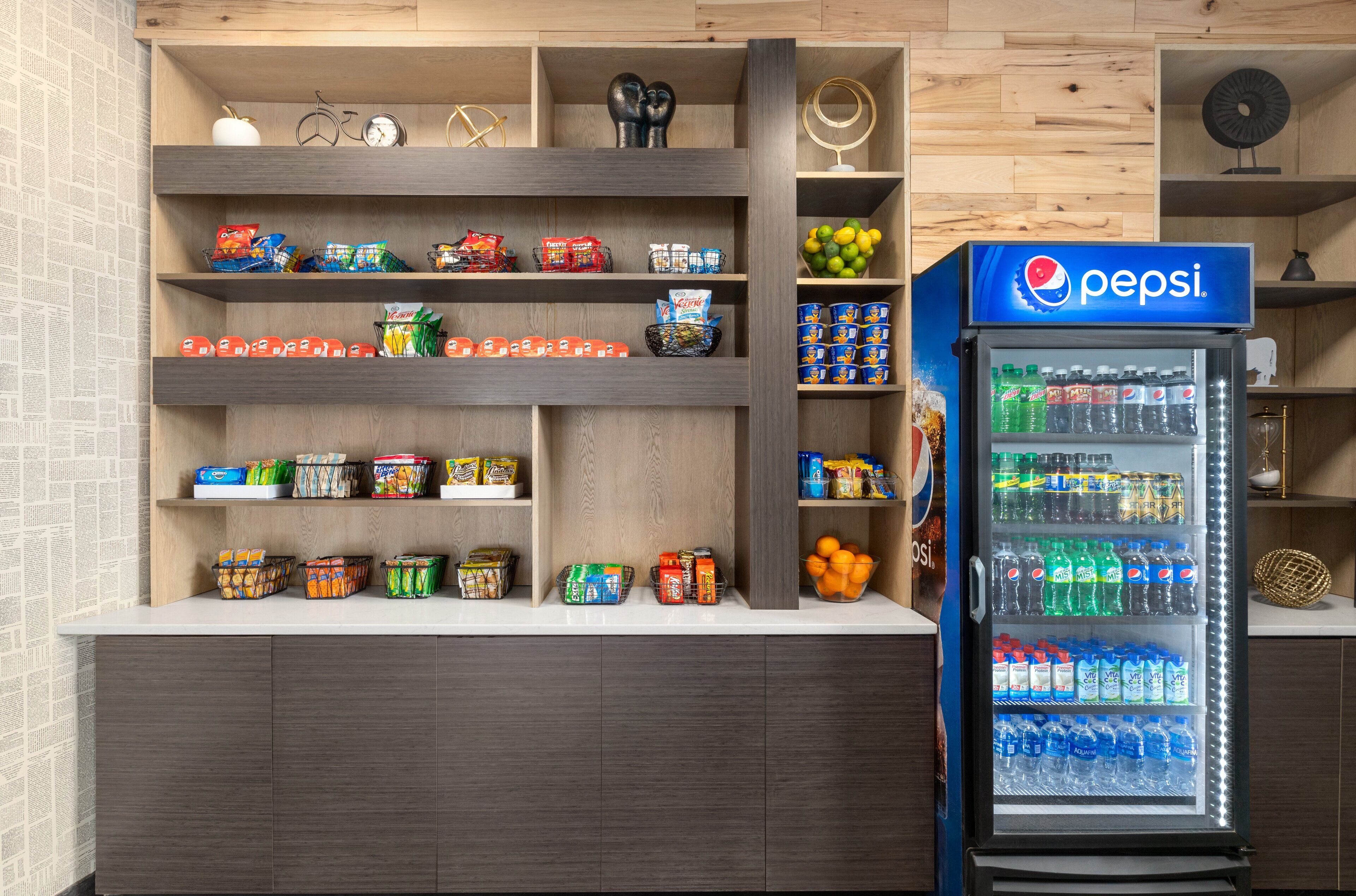 A kitchen with wooden shelves stocked with snacks, a refrigerator with Pepsi, and a clock on the wall.