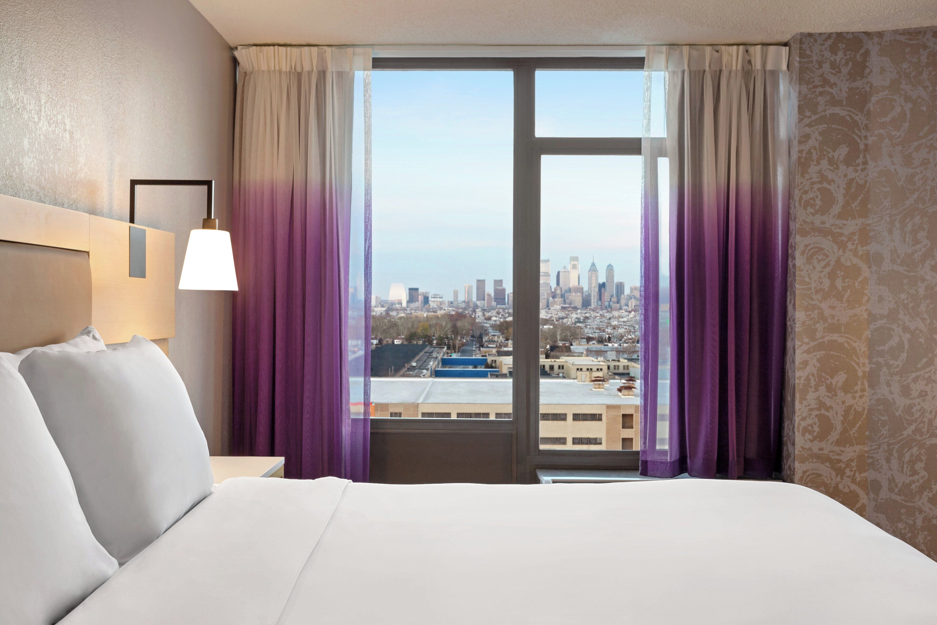Une chambre d’hôtel avec un grand lit, une table de chevet, une lampe et une vue sur le paysage urbain par une fenêtre ornée de rideaux violets.