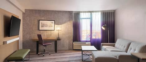 Habitación de hotel con sofá, escritorio y silla, televisor de pantalla plana, cuadro enmarcado, lámpara y ventana con cortinas.