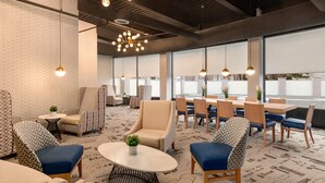 A modern lounge area with a mix of seating options, including sofas and chairs, arranged around a central table. The space features a patterned carpet, pendant lighting, and large windows offering views outside.